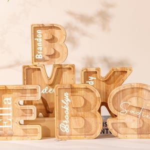 Könnte beinhalten: Eine Sammlung von fünf hölzernen buchstabenförmigen Spardosen mit durchsichtigen Acrylfronten. Die Buchstaben B, M, K, E und S sind mit weißen Vinylnamen wie Brandon, Ella, Brooklyn und Sadie personalisiert und haben eine natürliche Holzmaserung.