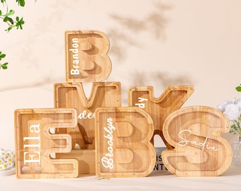 Peut inclure: Un ensemble de cinq tirelires en bois en forme de lettres avec des façades en acrylique transparent. Les lettres B, M, K, E et S sont personnalisées avec des prénoms en vinyle blanc comme Brandon, Ella, Brooklyn et Sadie, en bois naturel.