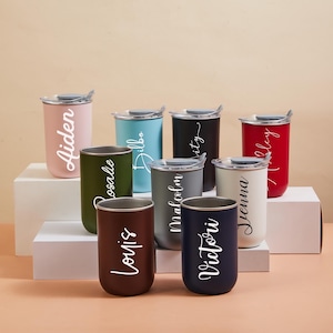 Könnte beinhalten: Eine Sammlung isolierter Becher in verschiedenen Farben, jeweils mit Deckel und einem Namen in Schreibschrift. Zu den Farben gehören Rosa, Blau, Schwarz, Rot, Grün, Grau, Braun und Weiß. Die Becher sind auf einer weißen Plattform angeordnet.