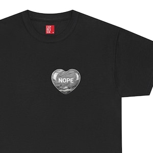 Puede incluir: Camiseta negra con un gráfico en forma de corazón que dice "NOPE" en texto blanco. El gráfico tiene una textura similar al mármol. En la esquina superior izquierda de la camiseta hay un pequeño cuadrado rojo con texto blanco.