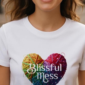 以下が含まれることがあります： 白いTシャツ。虹色の糸で作られたカラフルなハートのデザイン。ハートは虹色の糸で構成され、テキスト「Blissful Mess」がスクリプトフォントで下に印刷されています。
