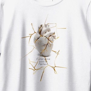 以下が含まれることがあります： 白いTシャツ。金色のひび割れ線が入った、様式化された解剖学的な心臓のデザイン。テキスト「Experience makes Perfect」が心臓の下にあります。デザインはシャツの中央に配置されています。