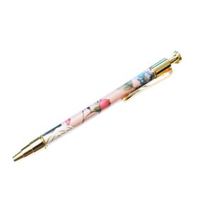 May include: A floral-patterned pen with a gold-colored clip and accents. The pen features a light pink background with a design of pink and blue flowers. The pen is a writing instrument.