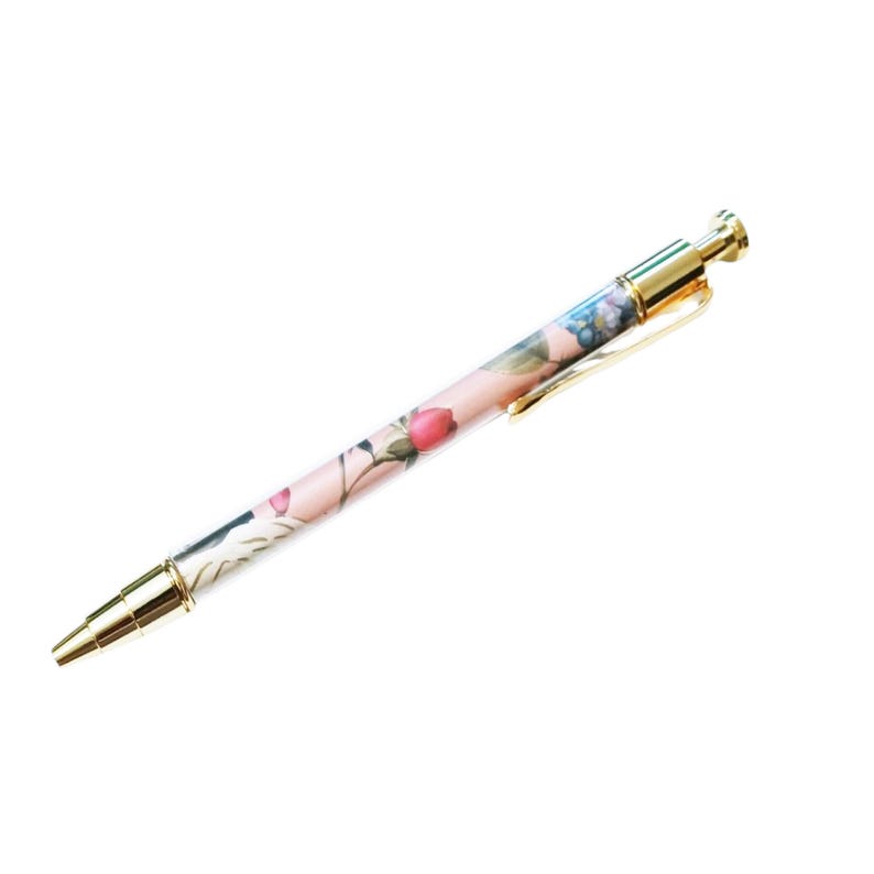 May include: A floral-patterned pen with a gold-colored clip and accents. The pen features a light pink background with a design of pink and blue flowers. The pen is a writing instrument.