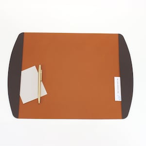 May include: A rectangular desk mat with rounded ends, featuring a burnt orange center and dark brown borders. A gold pen and a small stack of white notepaper are on the mat. The mat has a label that reads "San Lorenzo Intl, Inc."