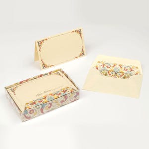 Peut inclure: Un ensemble de papeterie couleur crème avec une bordure décorative aux motifs floraux et volutes colorés. Le set comprend une carte pliée, une enveloppe assortie avec une doublure ornée et une boîte contenant des cartes avec le texte Kartos.