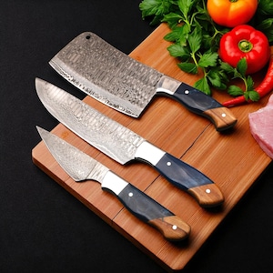 Könnte beinhalten: Set aus drei Küchenmessern aus Damaszener Stahl mit gemusterten Klingen und dunkelblauen und Holzgriffen. Die Messer liegen auf einem Holzschneidebrett, im Hintergrund frisches Gemüse.