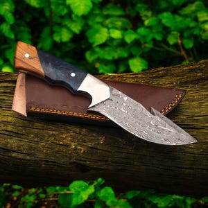 Könnte beinhalten: Ein Jagdmesser aus Damaststahl mit einem Holz- und dunkelblauen Griff, das auf einer braunen Lederscheide liegt. Die Klinge hat ein einzigartiges Muster. Das Messer wird auf einem Holzstamm mit grünem Laub im Hintergrund präsentiert.