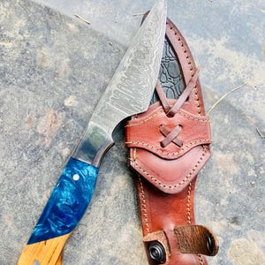 Könnte beinhalten: Ein handgefertigtes Messer mit einer gemusterten Stahlklinge und einem blau-holzfarbenen Griff. Das Messer wird mit einer braunen Lederscheide geliefert. Die Klinge ist ca. 15 cm lang.