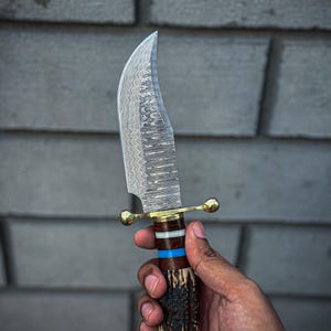 May include: A handcrafted hunting knife with a curved, patterned blade. The handle features a combination of dark brown, white, blue, and gold accents, with a textured, antler-like grip. The knife is held against a gray background.