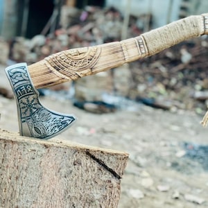 以下が含まれることがあります： 木製の柄と金属製の刃が付いた手作りの斧。柄は紐で巻かれ、彫刻が施されています。刃には複雑な彫刻が施されています。斧は木の表面に置かれています。