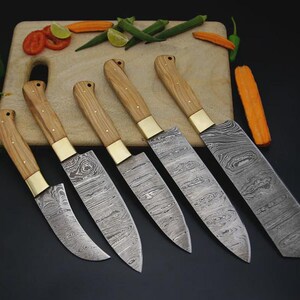 Könnte beinhalten: Set aus sechs Küchenmessern mit Holzgriffen und gemusterten Klingen. Die Messer haben Messingzwingen und sind auf einem Holzschneidebrett mit Gemüse angeordnet. Die Klingen weisen ein einzigartiges, wirbelndes Muster auf.