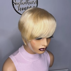 Könnte beinhalten: Blonde Kurzhaarperücke mit gestuftem Schnitt und seitlichem Pony. Sie wird auf einem Schaufensterkopf mit einem hellrosa ärmellosen Oberteil präsentiert. Im Hintergrund befindet sich ein Logo mit dem Text "Sogladhairs" in einem kreisförmigen Rahmen.