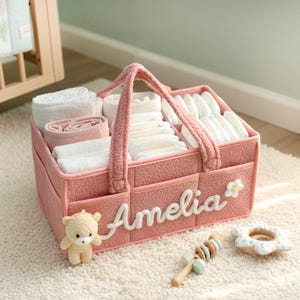Può includere: Un organizer per bambini in tessuto rosa con manico, riempito con pannolini, asciugamani e altri elementi essenziali per bambini. Il nome "Amelia" è visualizzato sul davanti. Un piccolo orsacchiotto, un sonaglio di legno e un anello per la dentizione sono accanto.