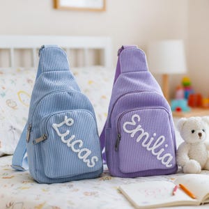 May include: Two corduroy sling bags, one light blue with "Lucas" and one lavender with "Emilia" embroidered in white. A white teddy bear and a notebook with crayons are also visible.
