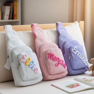 May include: Three corduroy sling bags in white, pink, and lavender, each with a front pocket and a name embroidered on the front. The names are "Ellie", "Megan", and "Emilia". A teddy bear and a book are also visible.