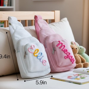 May include: Two corduroy sling bags, one white and one pink, each with a front pocket and embroidered name. The white bag has "Ellie" in colorful thread, and the pink bag has "Megan" in pink thread. The bags measure 9.4 inches high and 5.9 inches wide.