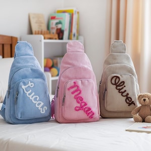 May include: Three corduroy sling backpacks in blue, pink, and beige, each with a front pocket and a name embroidered on the front. The names are Lucas, Megan, and Oliver. A teddy bear and a stuffed bunny sit nearby.