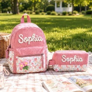 May include: A pink corduroy backpack and matching pencil case set, both featuring the name Sophia in white script lettering. The front pockets are decorated with a floral pattern and a small, three-dimensional crocheted flower applique.