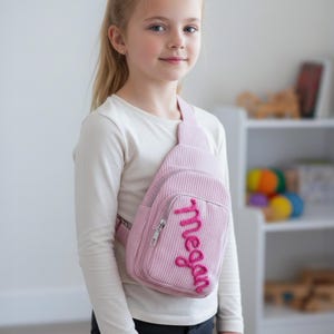 May include: A pink corduroy sling bag with a zipper pocket and the name "Megan" embroidered in a cursive font. The bag has a single strap and is worn across the body. The bag is designed for children.
