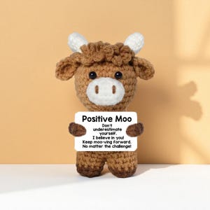 May include: A crocheted cow figurine in shades of brown, white, and tan, holding a sign that reads "Positive Moo." The sign includes motivational text. The cow has a textured appearance and is set against a solid background.