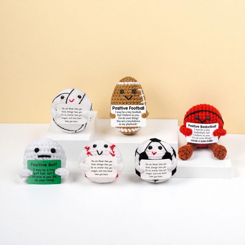 May include: A collection of crocheted sports balls, including a football, basketball, golf ball, and soccer ball, each with a smiling face and a motivational message. The balls are white, brown, red, and black.