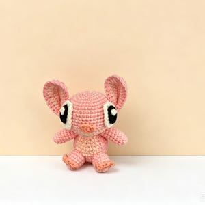Puede incluir: Una mu&ntilde;eca rosa de ganchillo hecha a mano con orejas grandes, ojos negros y blancos y brazos y piernas peque&ntilde;os. La mu&ntilde;eca est&aacute; hecha de hilo suave y tiene un aspecto texturizado. El fondo es de color melocot&oacute;n s&oacute;lido.