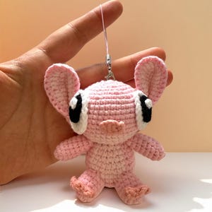 Puede incluir: Una mu&ntilde;eca rosa de ganchillo hecha a mano con orejas grandes, ojos negros y una nariz peque&ntilde;a. La mu&ntilde;eca tiene un cuerpo rosa claro y est&aacute; unida a una cuerda rosa con un cierre plateado. La mu&ntilde;eca est&aacute; en una mano.