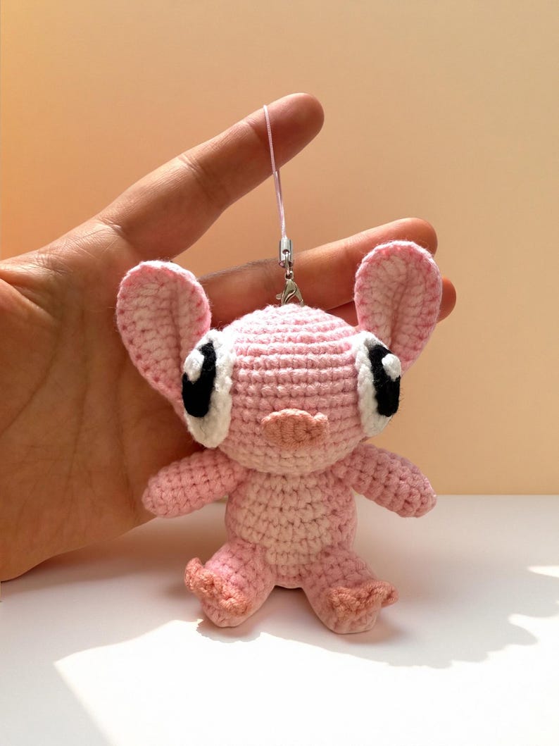 Puede incluir: Una mu&ntilde;eca rosa de ganchillo hecha a mano con orejas grandes, ojos negros y una nariz peque&ntilde;a. La mu&ntilde;eca tiene un cuerpo rosa claro y est&aacute; unida a una cuerda rosa con un cierre plateado. La mu&ntilde;eca est&aacute; en una mano.