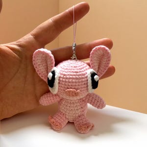 Puede incluir: Una mu&ntilde;eca rosa de ganchillo hecha a mano con orejas grandes, ojos negros y blancos y una peque&ntilde;a nariz rosa. La mu&ntilde;eca tiene un cuerpo peque&ntilde;o y est&aacute; suspendida por un hilo rosa. La mu&ntilde;eca es un personaje lindo y caprichoso.