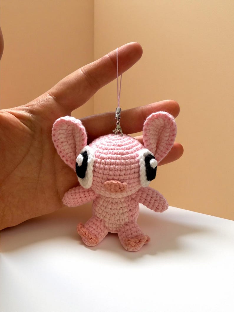 Puede incluir: Una mu&ntilde;eca rosa de ganchillo hecha a mano con orejas grandes, ojos negros y blancos y una peque&ntilde;a nariz rosa. La mu&ntilde;eca tiene un cuerpo peque&ntilde;o y est&aacute; suspendida por un hilo rosa. La mu&ntilde;eca es un personaje lindo y caprichoso.