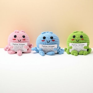 May include: Three crocheted octopus toys in pink, blue, and green. Each octopus holds a small white card with the text "Positive Octopus" and an inspirational message. The toys have black button eyes and smiling faces.