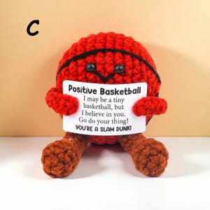 May include: A handmade, red crocheted basketball with black stripes, holding a sign that reads "Positive Basketball." The sign includes an inspirational message. The basketball has brown crocheted legs and arms, and black button eyes.
