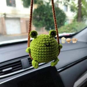 May include: A handmade green crochet frog car charm hanging from a brown string. The frog has large eyes and small arms and legs. The charm is suspended in front of a car's dashboard.
