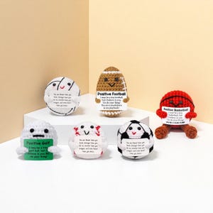 May include: A selection of handmade crochet sports balls, including a golf ball, baseball, soccer ball, football, and basketball. Each ball has a smiling face and a motivational message on a small sign.