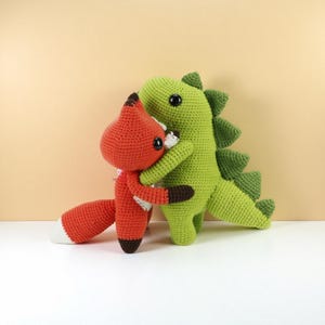 May include: Two crocheted stuffed animals: a red fox and a green dinosaur, embracing. The fox has a white chest and tail tip, with brown accents. The dinosaur has a spiky back. Both have black eyes and are made of yarn.