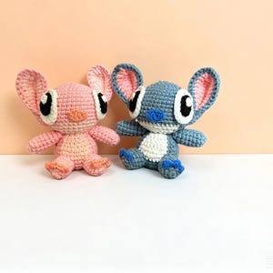 Puede incluir: Dos peluches de ganchillo, uno rosa y otro azul, que se asemejan a personajes de dibujos animados. El juguete rosa tiene orejas rosas con un interior rosa claro, y el juguete azul tiene orejas azules con un interior rosa. Ambos tienen ojos negros grandes y est&aacute;n sentados.