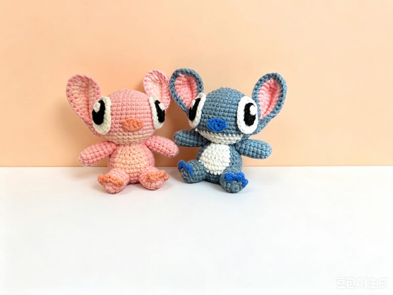 Puede incluir: Dos peluches de ganchillo, uno rosa y otro azul, que se asemejan a personajes de dibujos animados. El juguete rosa tiene orejas rosas con un interior rosa claro, y el juguete azul tiene orejas azules con un interior rosa. Ambos tienen ojos negros grandes y est&aacute;n sentados.