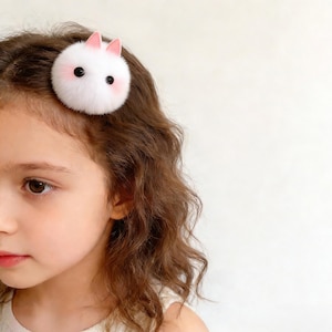 May include: A white and pink bunny hair clip. The clip features a fluffy white body with pink cheeks, ears, and black eyes. The hair accessory is attached to a child's brown hair.