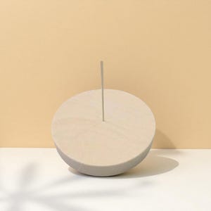 May include: A light beige incense holder with a circular base and a single, thin, gray incense stick. The holder is made of wood and has a smooth, matte finish. The background is a soft, warm beige.
