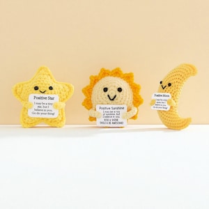 Pode incluir: Três figuras amarelas de crochê: uma estrela, um sol e uma lua crescente. Cada figura segura uma pequena placa branca com mensagens positivas. A estrela, o sol e a lua têm rostos sorridentes. O fundo é amarelo claro liso.