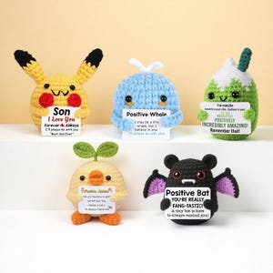 May include: A collection of five crocheted amigurumi figures, each holding a small sign with positive messages. The figures include a yellow Pikachu-like character, a blue whale, a green matcha-like character, a yellow chick, and a black bat. The signs have encouraging phrases.