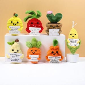 Puede incluir: Una colección de peluches de ganchillo hechos a mano con forma de frutas y verduras, como un limón, un rábano, un cactus, una pera, una zanahoria, una naranja y un maíz. Cada peluche sostiene un pequeño cartel con un mensaje inspirador.