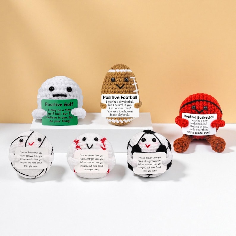 May include: A collection of crocheted sports balls, including golf, football, basketball, volleyball, and soccer. Each ball has a smiling face and a small sign with an inspirational message. The balls are white, brown, red, and black.
