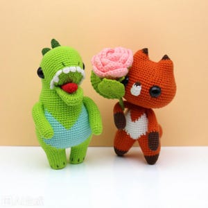 May include: Two handmade crochet amigurumi figures: a green dinosaur with a blue belly and a red tongue, and an orange fox holding a pink flower. The figures have black eyes and are made with yarn.