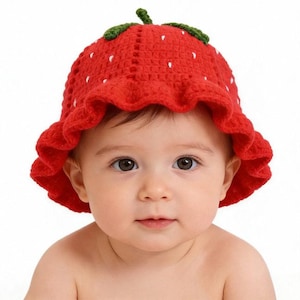 May include: A red crochet baby hat shaped like a strawberry, with white seed details and a green stem. The hat has a ruffled brim and is worn by a baby with brown eyes.