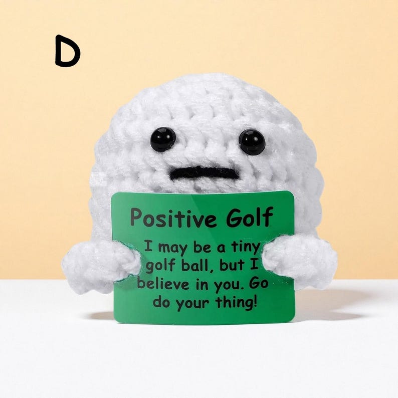 May include: A white crocheted golf ball character with black button eyes and a stitched mouth. It holds a green sign that reads "Positive Golf" and a motivational message. The letter "D" is in the upper left corner.