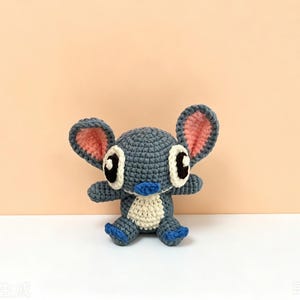 Puede incluir: Un juguete de peluche de ganchillo hecho a mano con forma de personaje de dibujos animados. El juguete es principalmente azul con orejas internas rosas, detalles blancos y ojos negros. Tiene brazos y piernas peque&ntilde;os y est&aacute; sentado.
