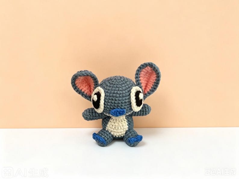 Puede incluir: Un juguete de peluche de ganchillo hecho a mano con forma de personaje de dibujos animados. El juguete es principalmente azul con orejas internas rosas, detalles blancos y ojos negros. Tiene brazos y piernas peque&ntilde;os y est&aacute; sentado.