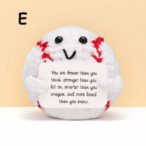 May include: A white crocheted baseball with red stitching and black eyes, holding a card with the text "You are braver than you think, stronger than you let on, smarter than you imagine, and more loved than you know."